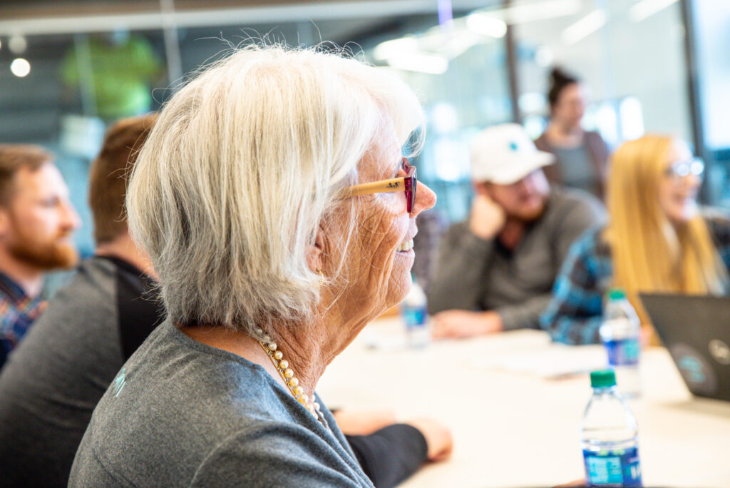 Woman with short silver hair is in focus with a conference room full of people behind her out of focus