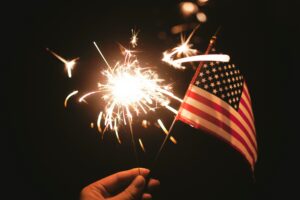 A hand holds a sparkler in front of an american flag at night time