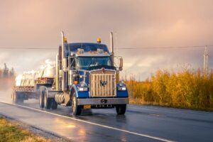 A blue semi truck drives down a wet road with the sun setting behind it